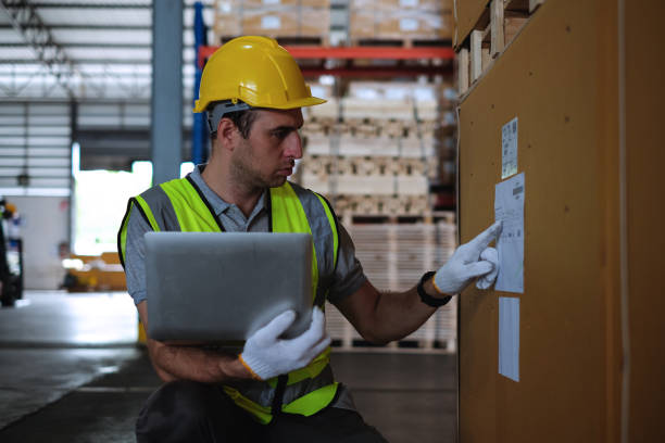 Caucasian male engineer wearing safety helmet and vest holding laptop and looking part number from product box at automobile factory.