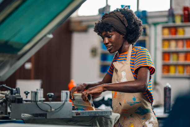 Multicultural female graphic technologist using rubber blade for silk screen printing at printing workshop. Smiling african american printing specialist using squeegee for screen printing on a press.
