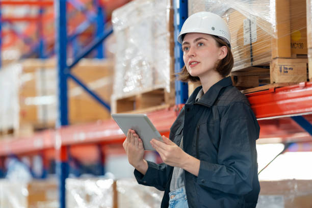 Warehouse quality inspector improve logistic and shipping performance. Caucasian warehouse manager woman with safety helmet holding tablet looking at shelf rack in the distribution center. Check inventory stock from tablet. Logistic and export goods with global business